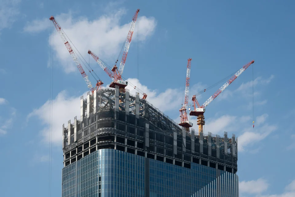 A glass tower under construction, with cranes at its top